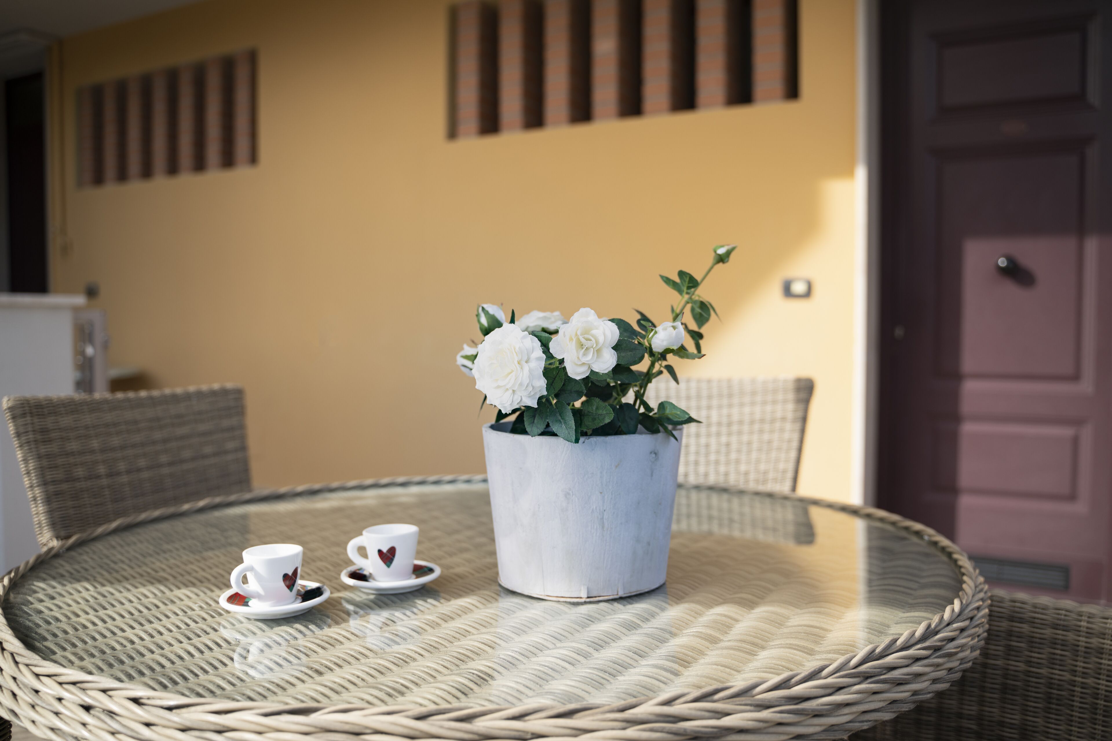 A round wicker table with a glass top, two white cups with heart designs, and a potted white flower arrangement.