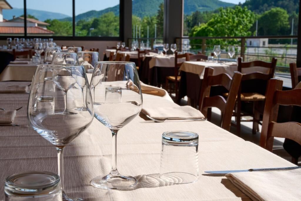 Un ristorante con tavoli apparecchiati, bicchieri trasparenti e vista su spazi verdi e montagne.
