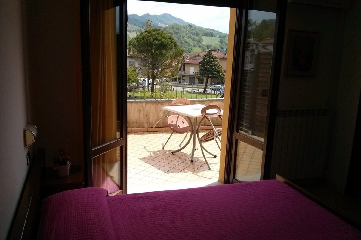 A room with a bed, a window, and a view of a balcony with a table and chairs.