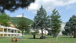 A grassy area with a building in the background, a bench, and a playground structure. - Shine (Acqualagna)
