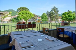 An outdoor dining area with tables set for two, a view of a playground, and a grassy field with trees. - Shine (Acqualagna)