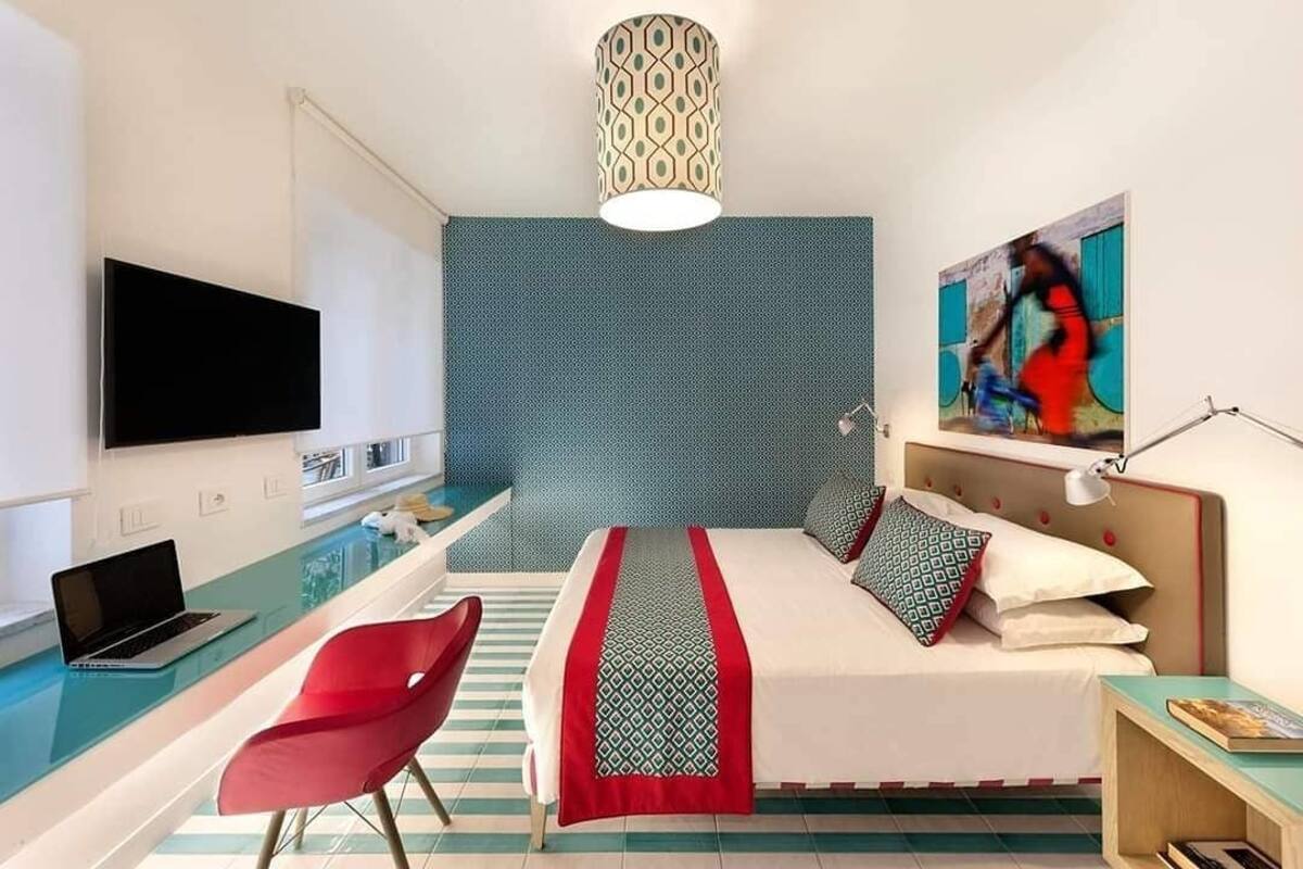 Een hotelkamer met een bed, een bureau met een laptop, een rode stoel, een televisie en een schilderij aan de muur.