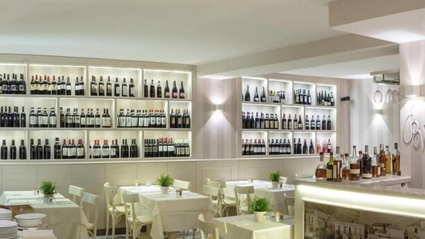 L'intérieur d'un restaurant, avec de nombreuses tables, des chaises et un mur dédié à l'exposition des bouteilles de vin.