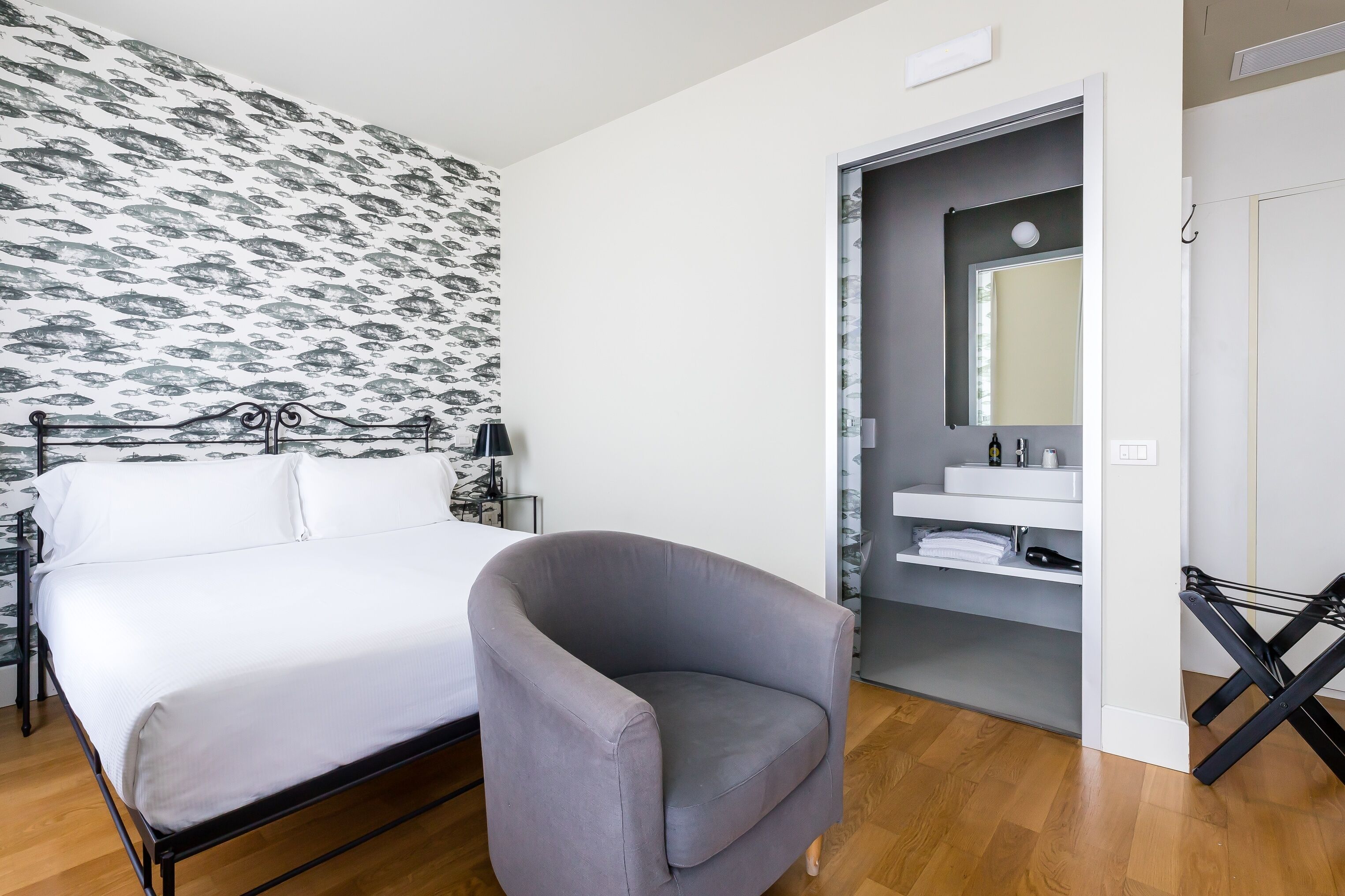 A bedroom with a bed, a grey armchair, a patterned wallpaper, a bathroom with a mirror, and a shelf with towels.