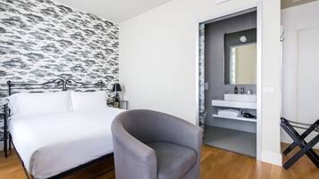 A bedroom with a bed, a grey armchair, a patterned wallpaper, a bathroom with a mirror, and a shelf with towels.