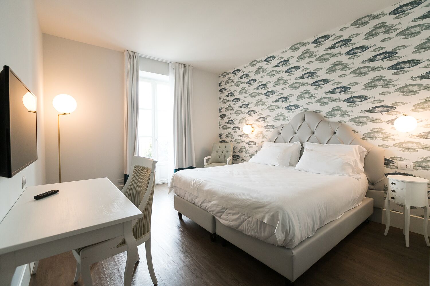 A hotel room with a bed, a desk, a chair, a TV, and a wall with a fish pattern.