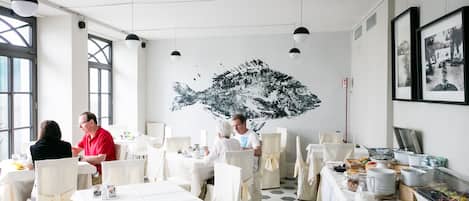 A restaurant with white tables and chairs, a large abstract painting on the wall, and framed pictures on the wall.
