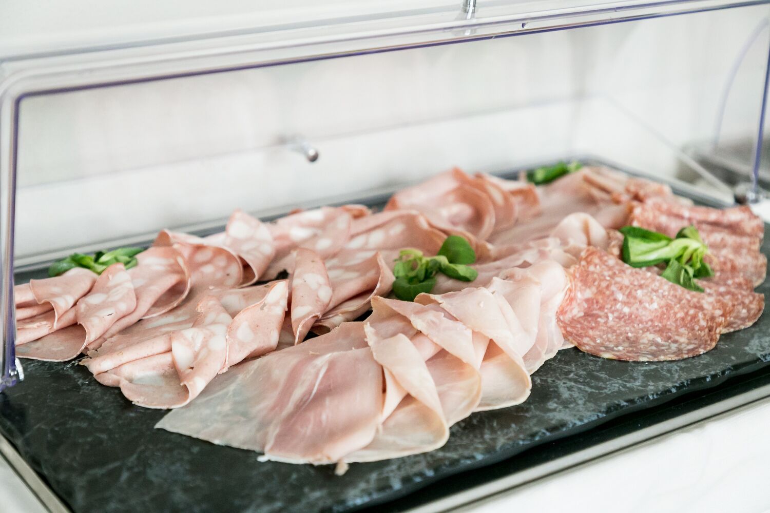 A display of various meats, including sliced salami and prosciutto, arranged on a black slate surface.