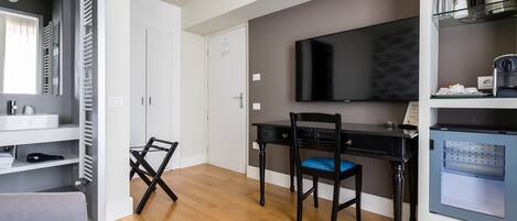 A modern hotel room with a wooden floor, a desk with a chair, a flat-screen TV, a built-in mini-fridge, and a small kitchenette.