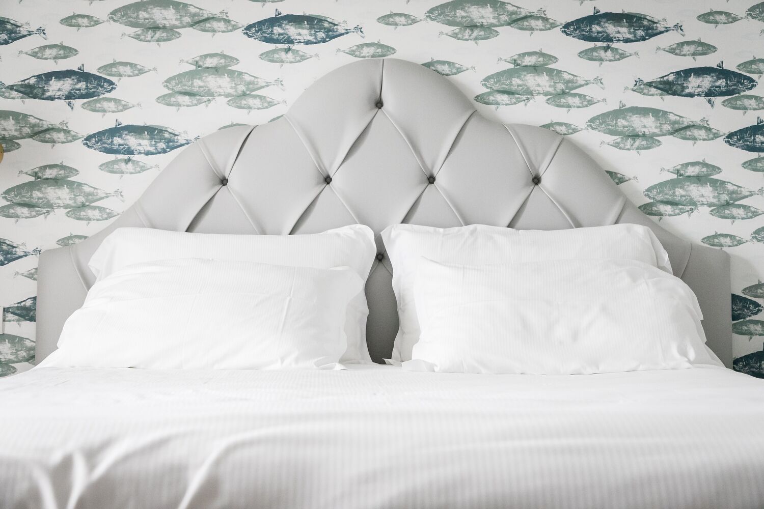 Una cama con cabecera capitonada y ropa de cama blanca frente a un papel tapiz con motivos de peces.