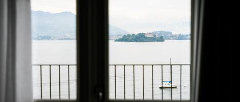 A view through a window with a railing, overlooking a calm body of water with a small boat and distant land with buildings.
