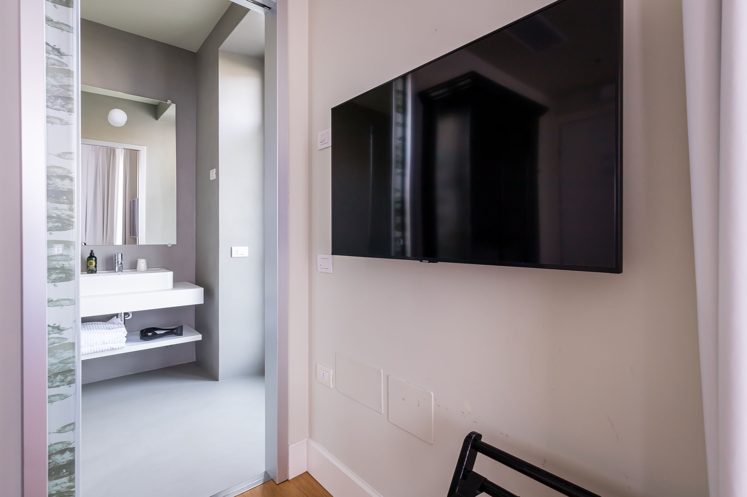 A modern bathroom with a white sink, mirror, and towel rack. A black television is mounted on the wall.