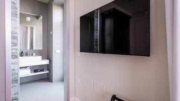 Un baño moderno con un lavabo blanco, espejo y toallero. Un televisor negro está montado en la pared.