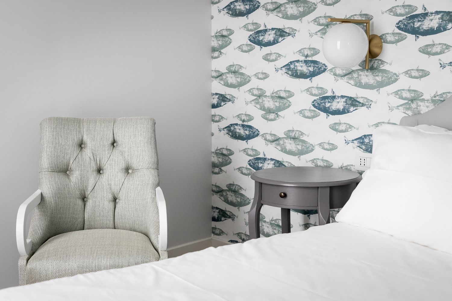 A bedroom with a tufted armchair, a bedside table, and a wall with a fish pattern wallpaper.