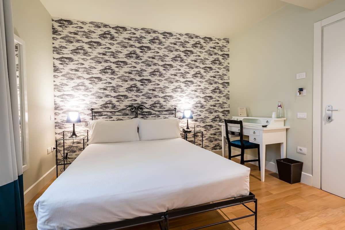 A bedroom with a bed, two bedside lamps, a desk with a chair, and a small trash bin.