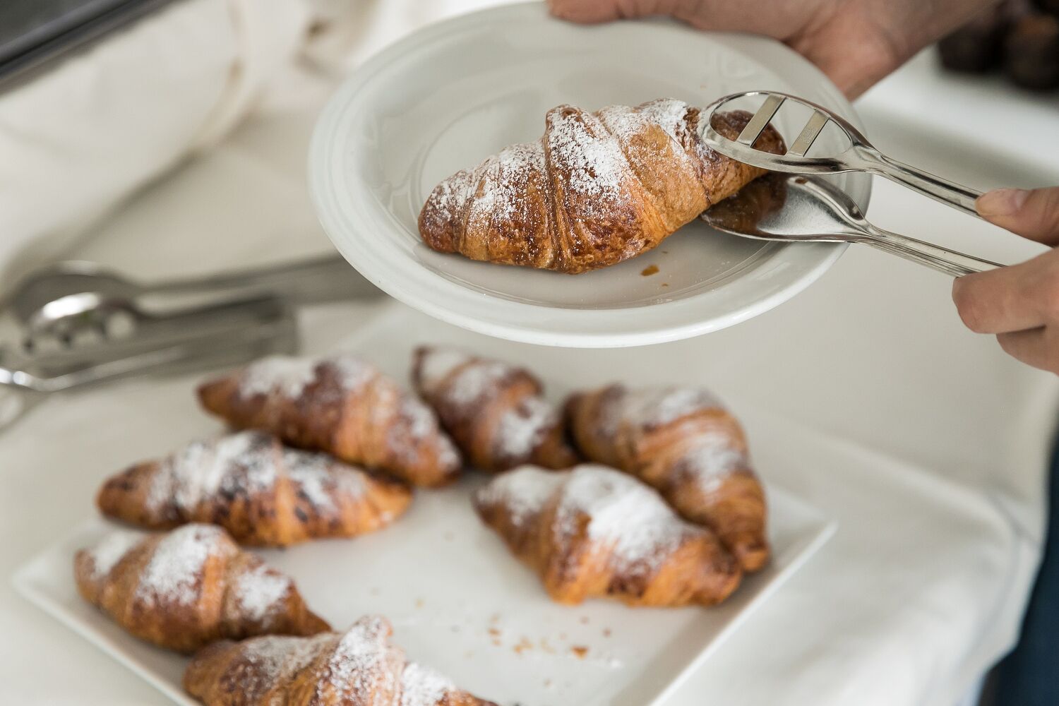 A person holding a plate with a croissant and tongs, with more croissants on a plate in the background.