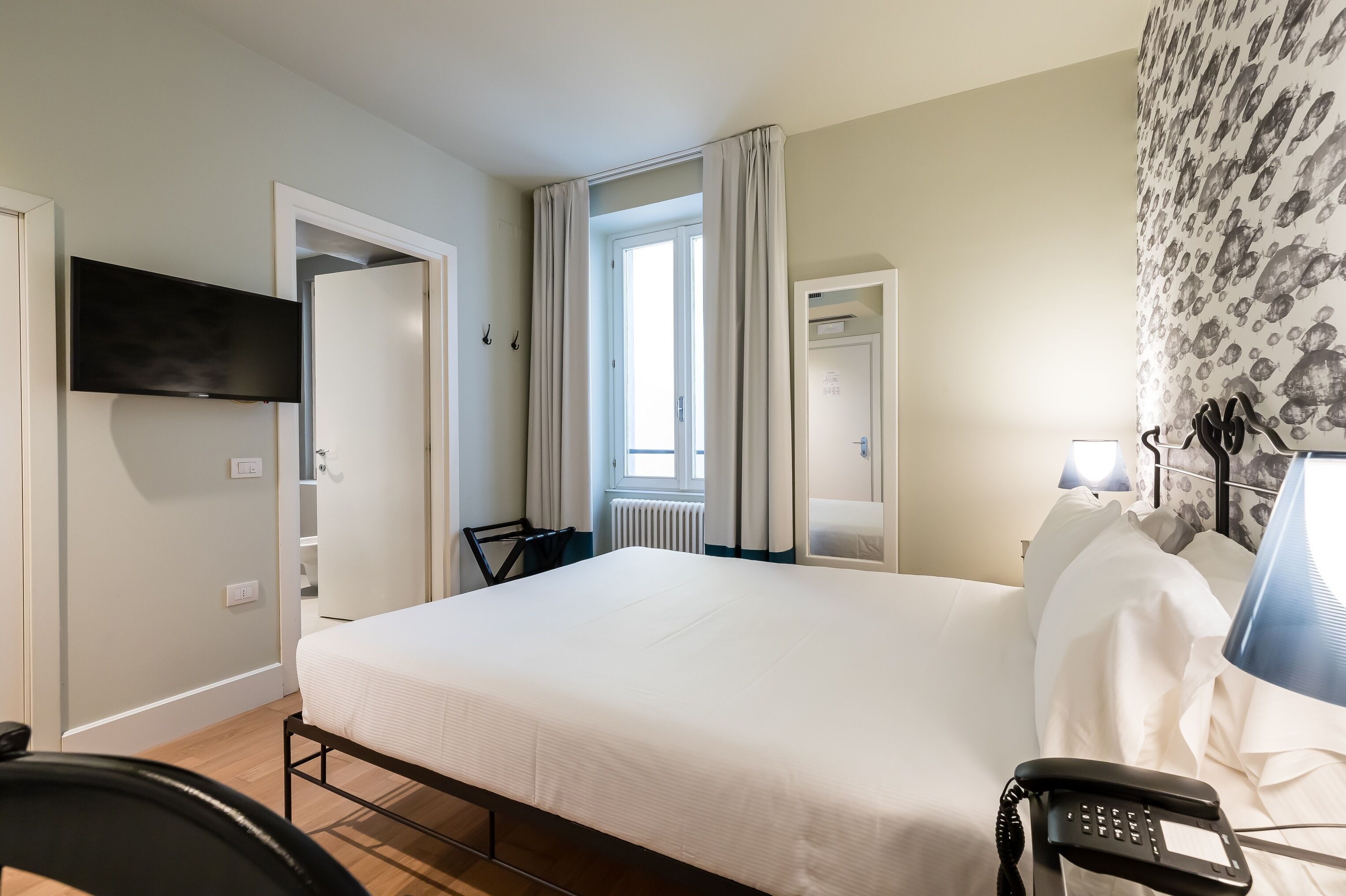 A hotel room with a bed, a desk, a chair, a telephone, a lamp, and a window with curtains.
