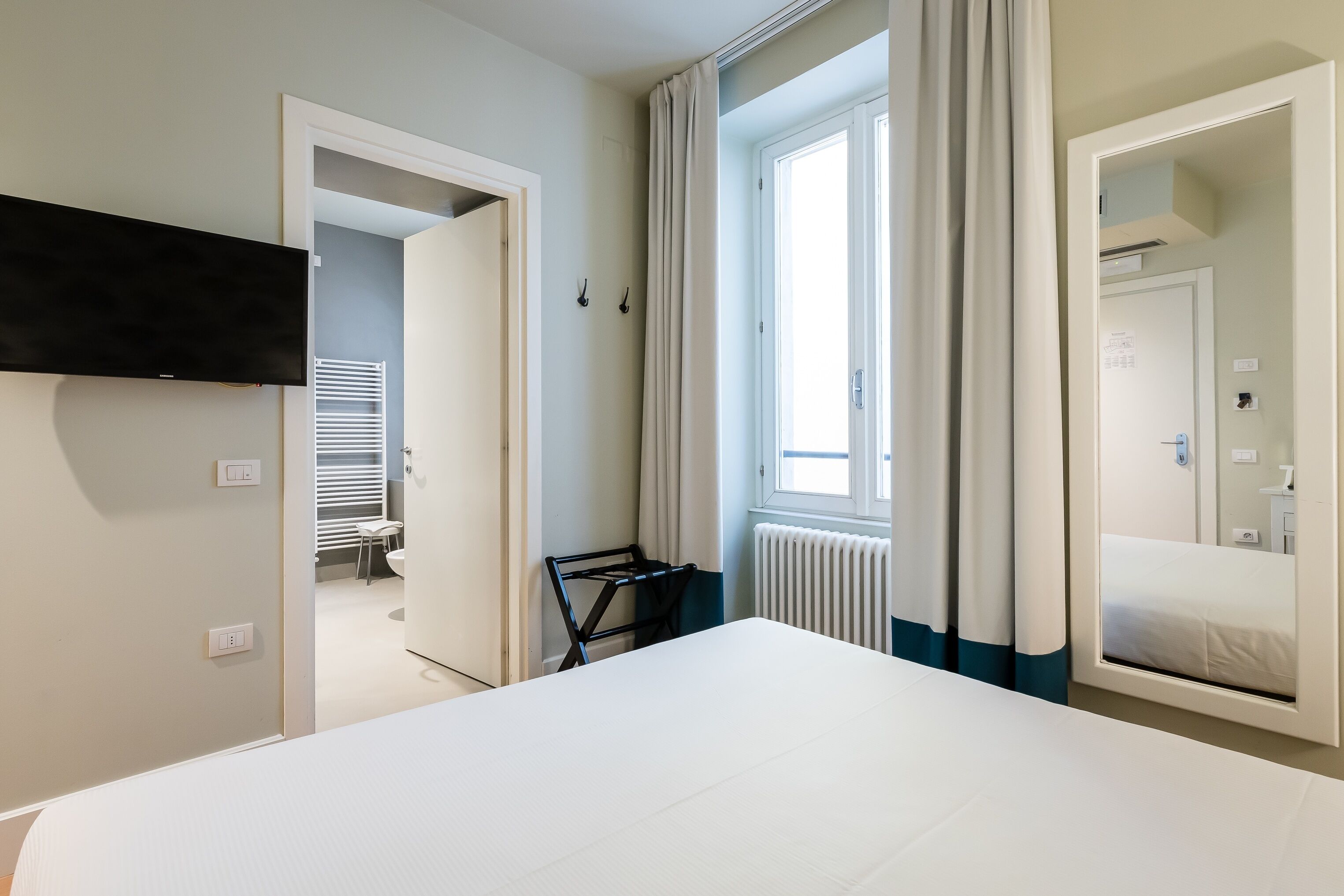 A modern hotel room with a large bed, a flat-screen TV mounted on the wall, a radiator, and a window with curtains.