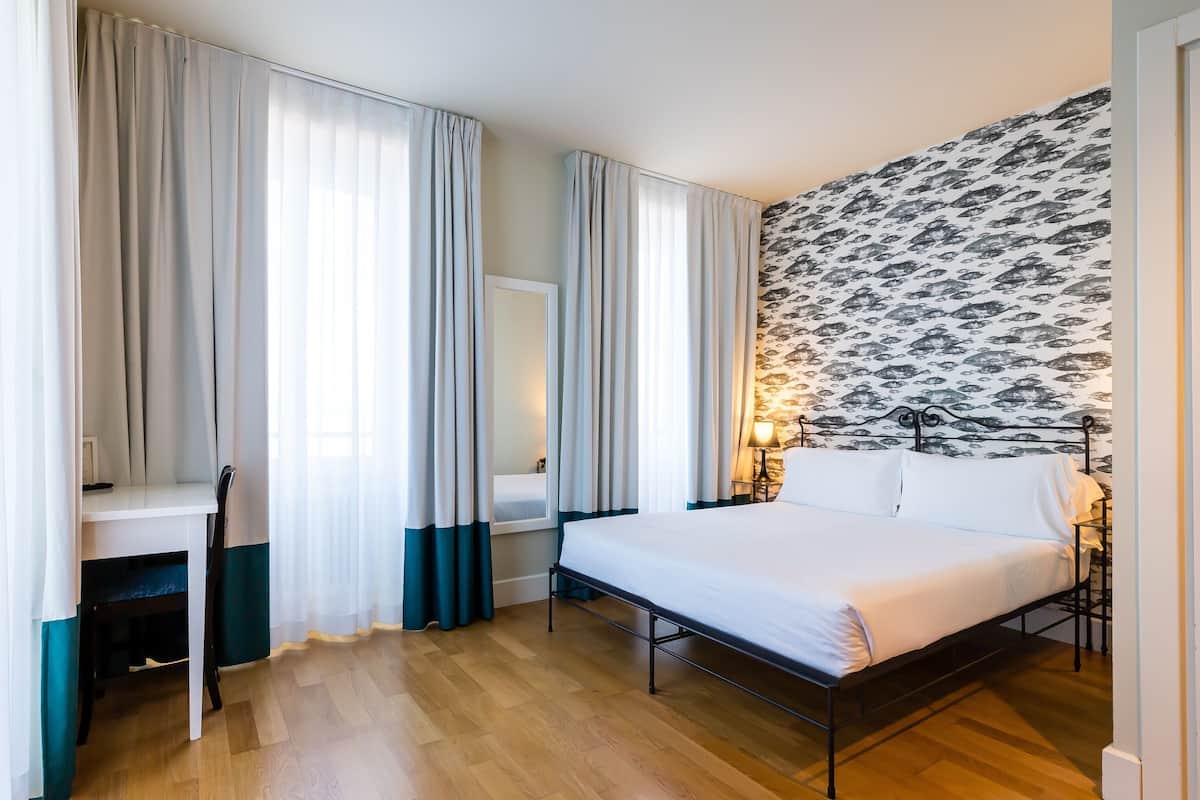 A hotel room with a bed, a desk with a chair, a mirror, and a patterned wall.