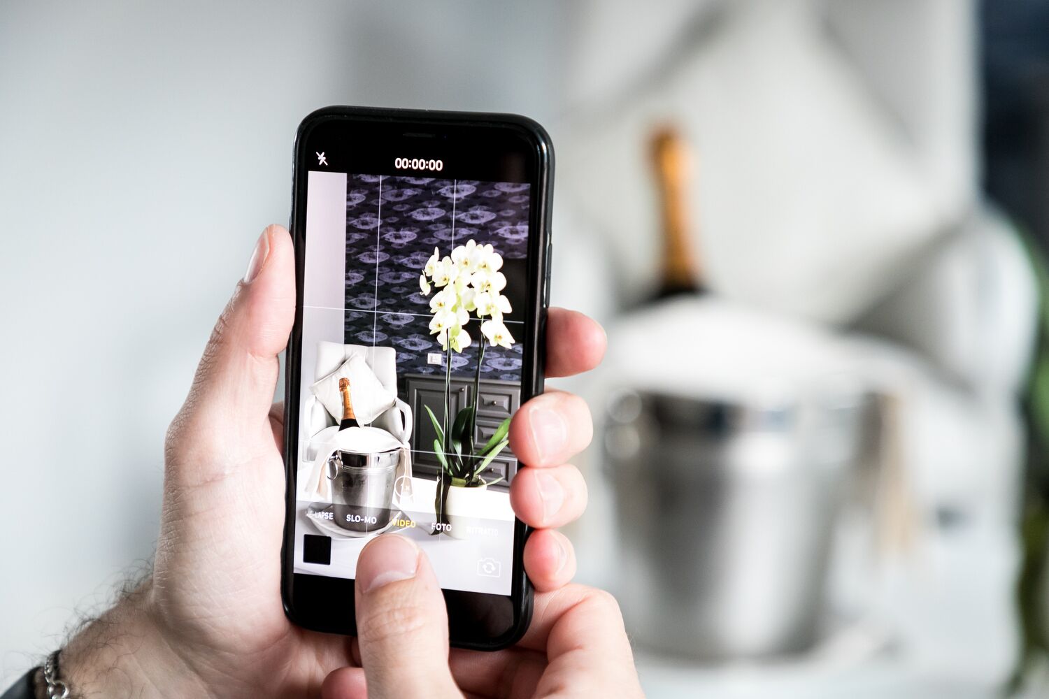 A hand holding a smartphone displaying a photo of a champagne bottle, a flower, and a pot.