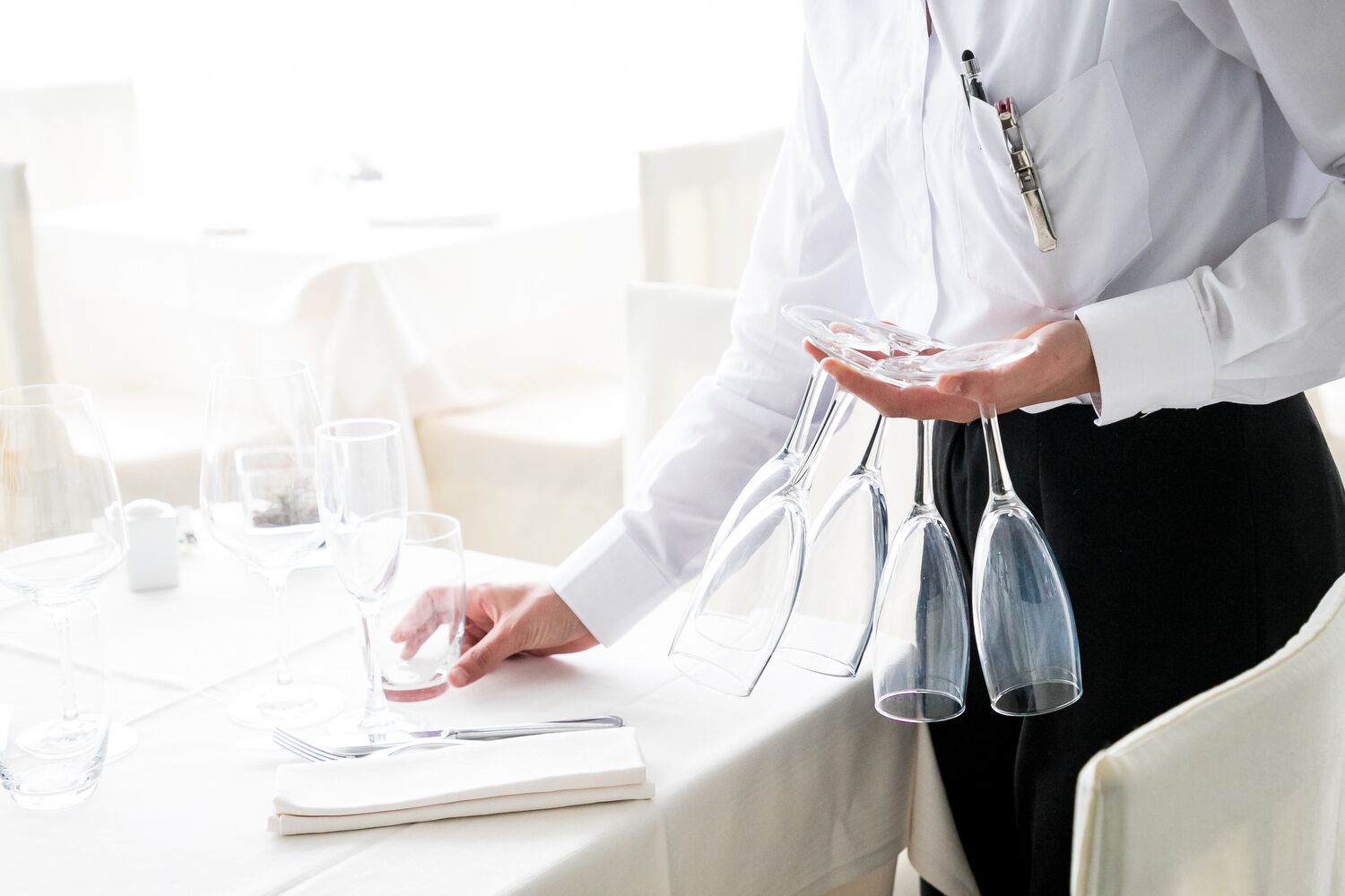 A person holding wine glasses on a table with a white tablecloth, glasses, and a notebook.