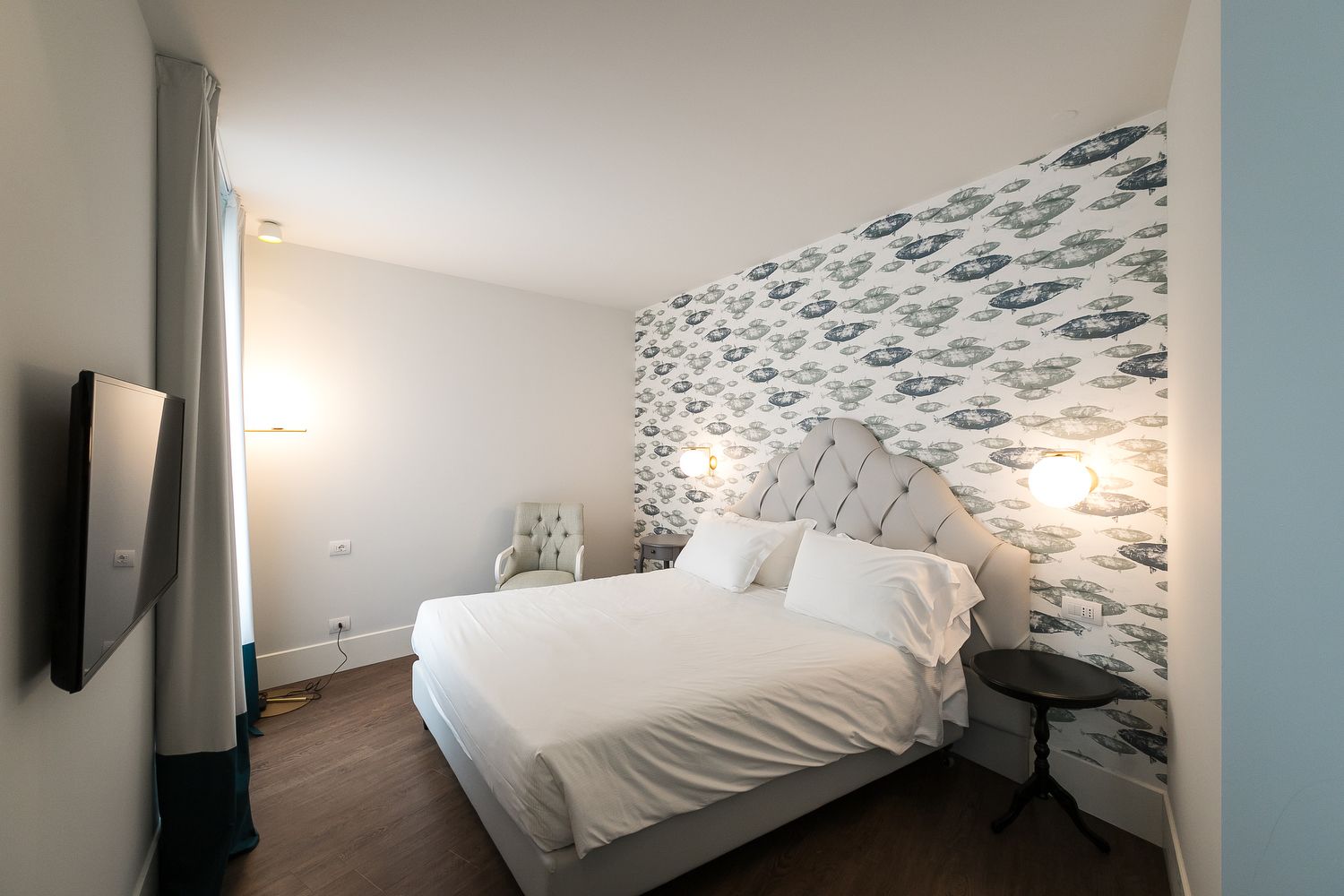 Un dormitorio con cama, una silla, mesita de noche y televisor montado en la pared.