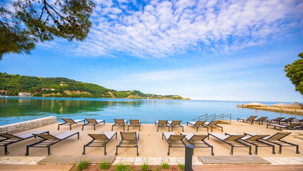 Una terraza con sillones de sol con vistas a una masa de agua tranquila, una terraza de madera y un área ajardinada con plantas.