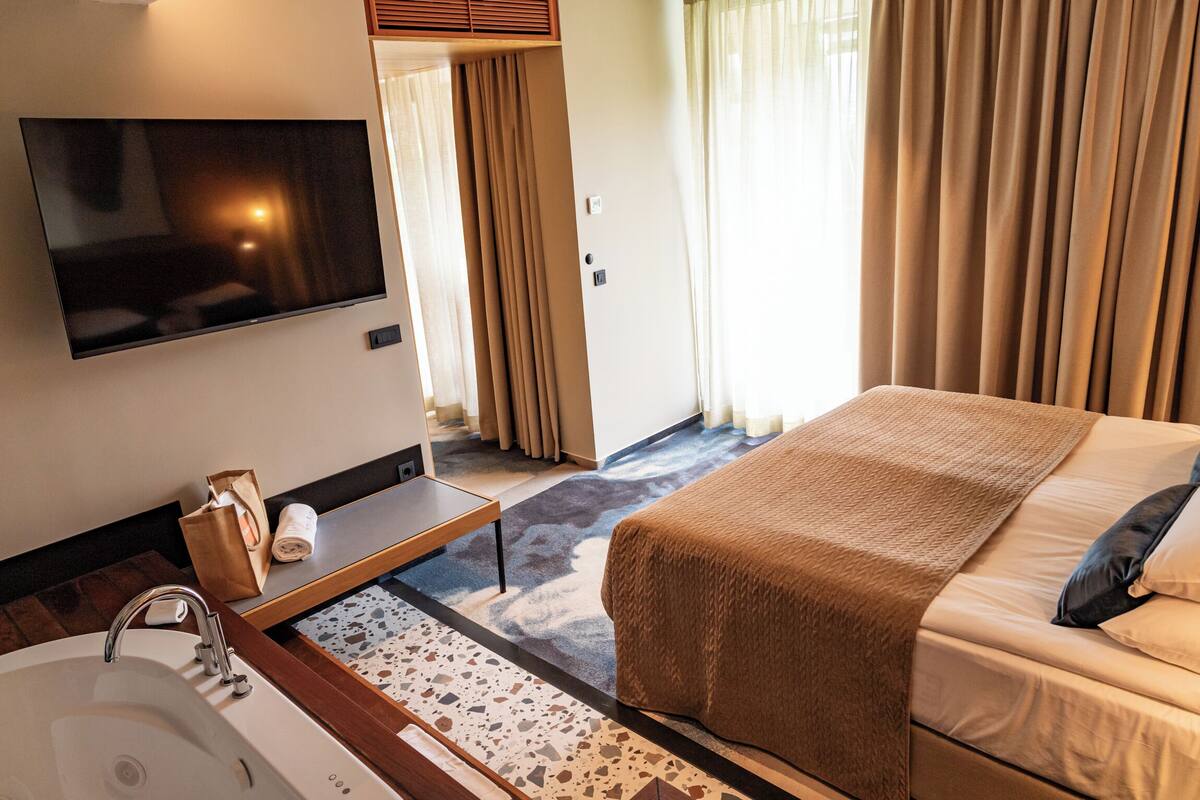 Une chambre d’hôtel avec un lit, un téléviseur fixé au mur, une table de chevet avec une bouteille et une boîte de mouchoirs, une salle de bain avec un lavabo et un miroir, et une fenêtre avec des rideaux.