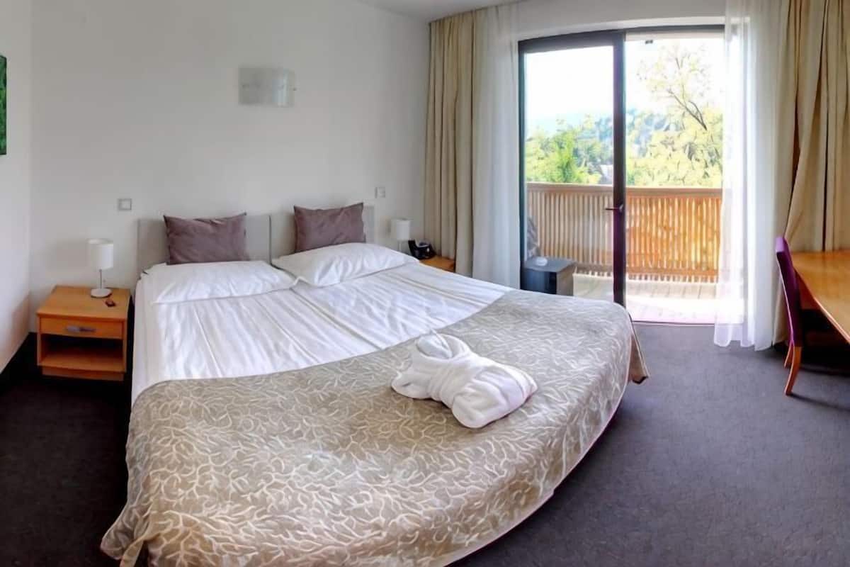Habitación de hotel con una cama grande, un escritorio, una silla, una mesita de noche y un balcón con vistas.