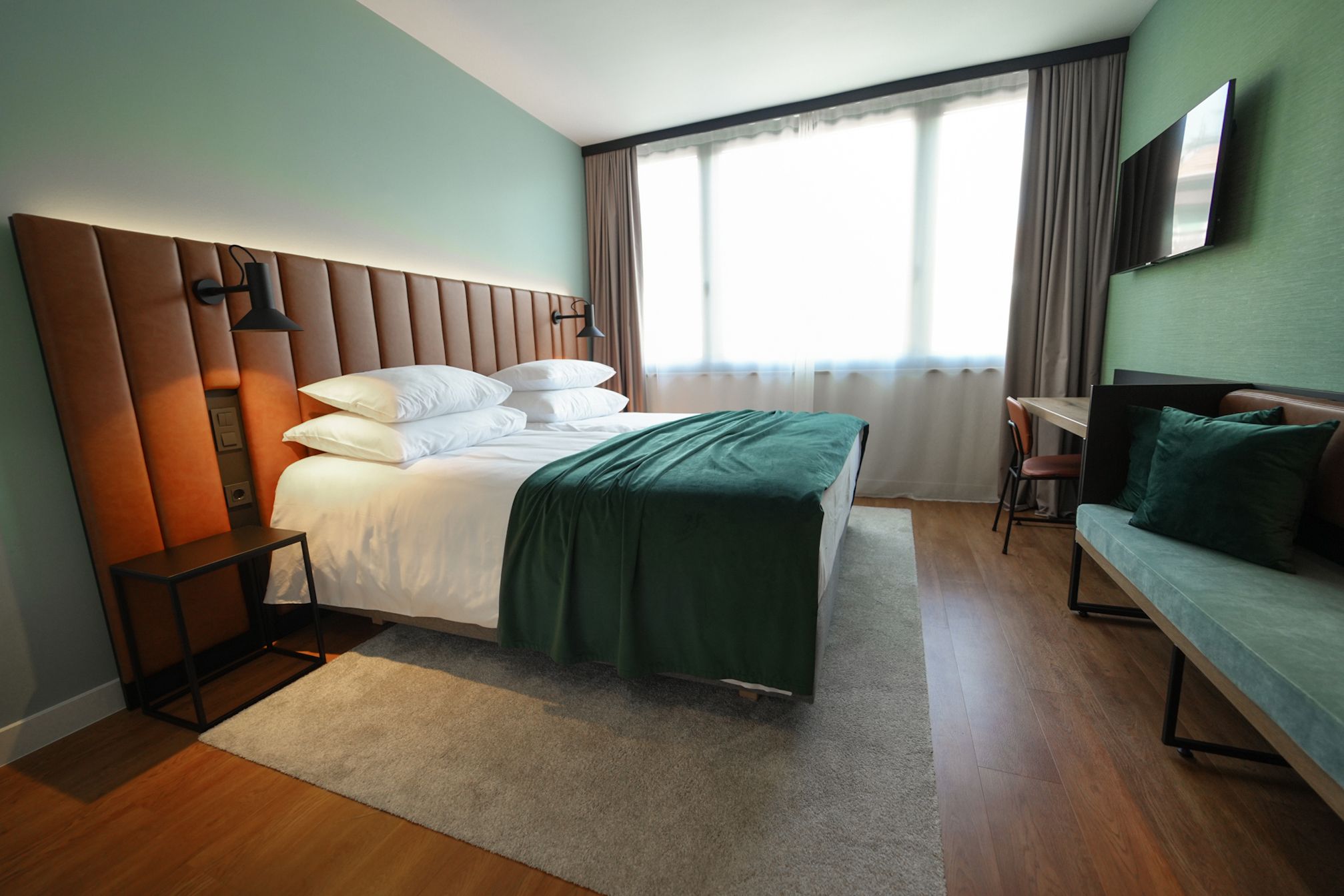 A hotel room with a bed, a desk, a chair, a sofa, a TV, and a window with curtains.