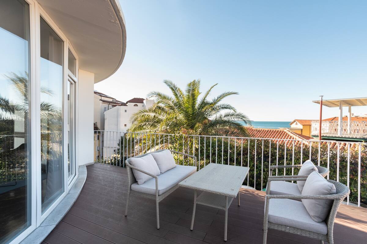 A balcony with wicker furniture, a view of the sea, and a palm tree.