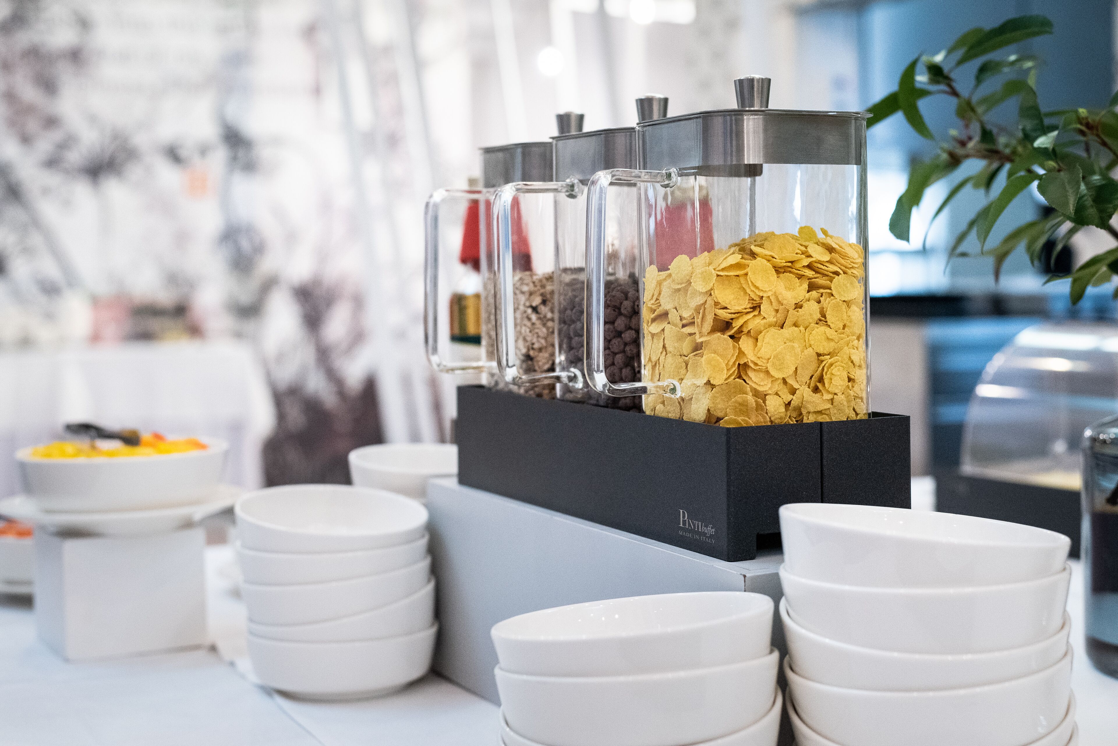Montaje de bufé con recipientes de vidrio transparente que contienen diversos alimentos, platos blancos apilados sobre una mesa y una caja negra con la etiqueta 'PANTHEA'.