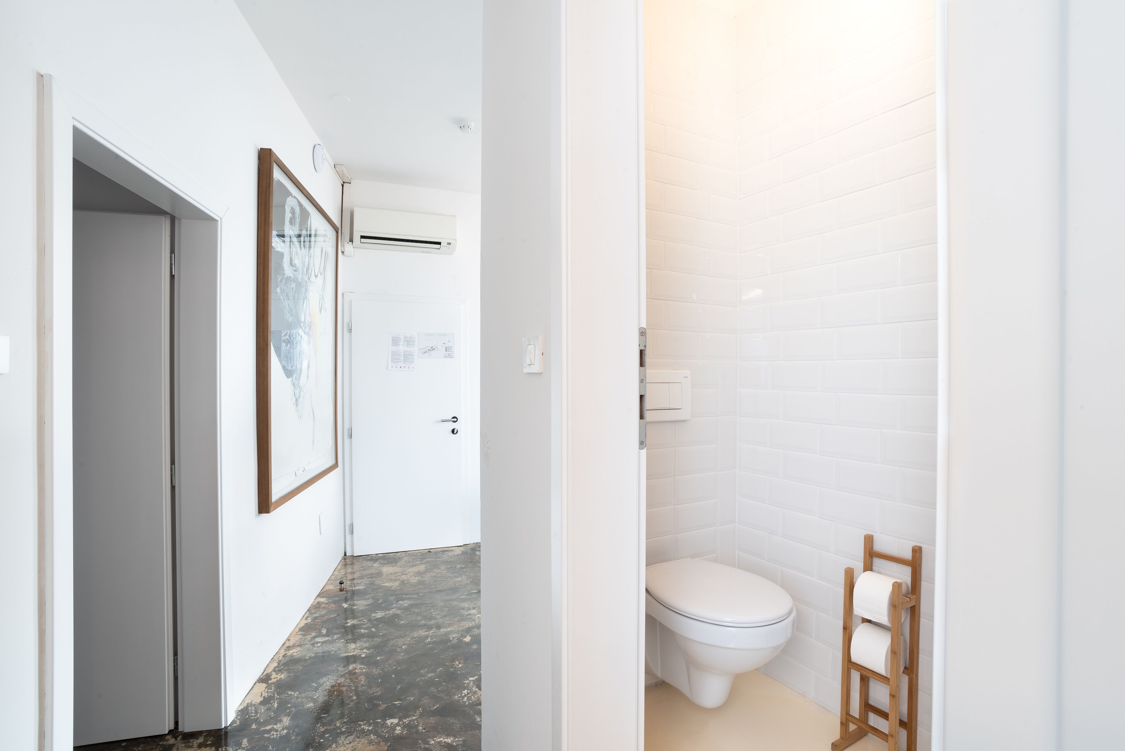 Pasillo que lleva a un baño con azulejos blancos, un inodoro y un porta papel higiénico de madera.