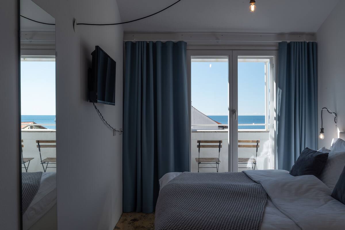A hotel room with a bed, a TV mounted on the wall, and a view of the sea through the windows.