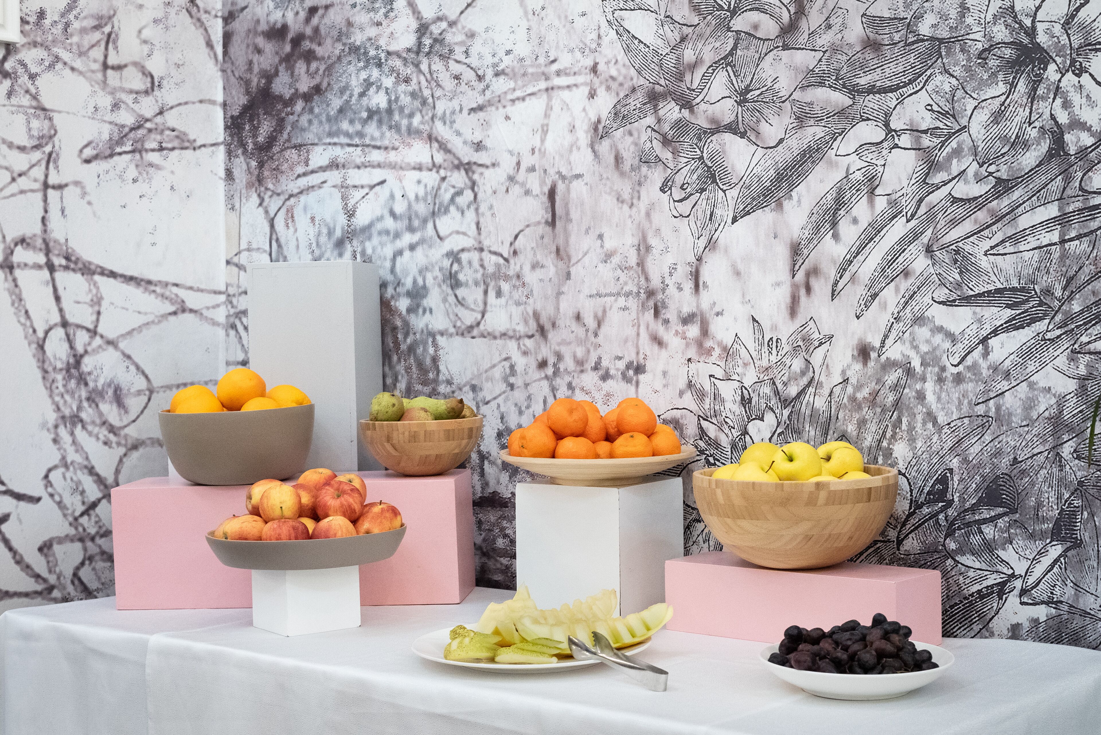 A table with various fruits and bowls of oranges, apples, and grapes, set against a textured wall.