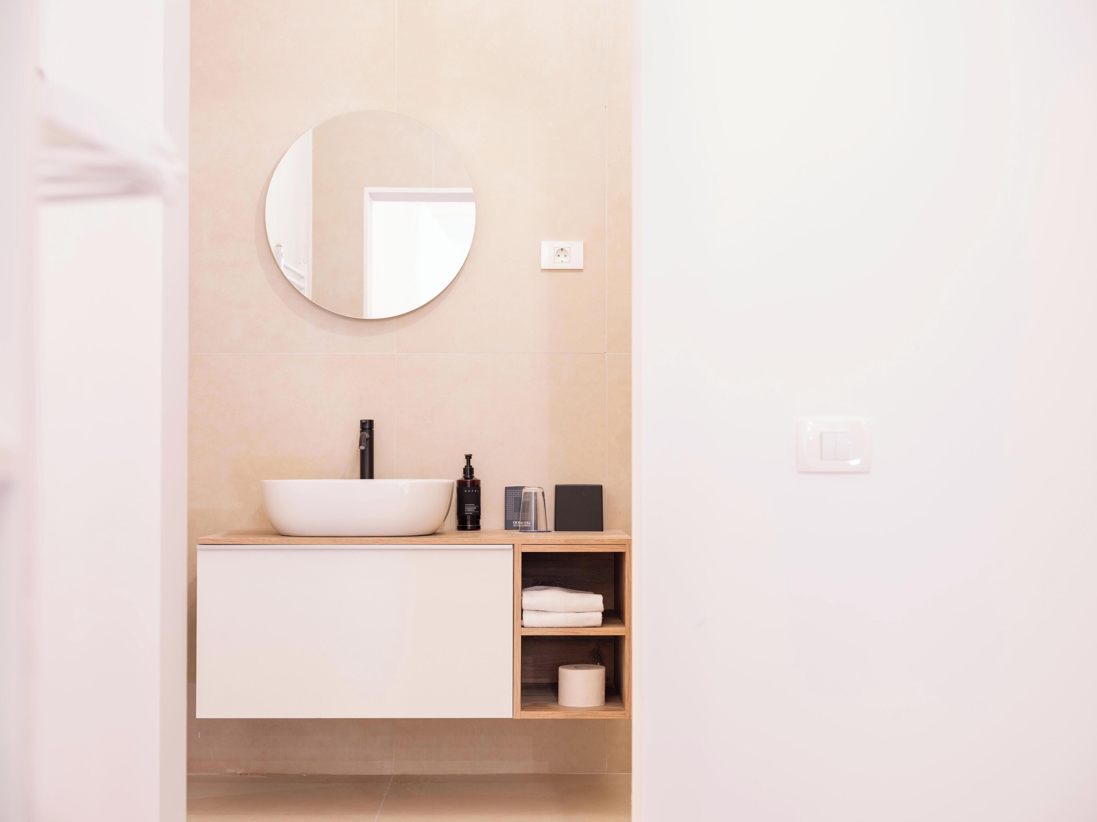 Un bagno moderno con un lavandino bianco, uno specchio rotondo e uno scaffale con asciugamani e prodotti per l'igiene personale.