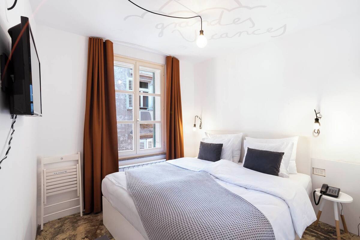 A hotel room with a bed, a bedside table with a phone, a window with curtains, and a wall-mounted TV.