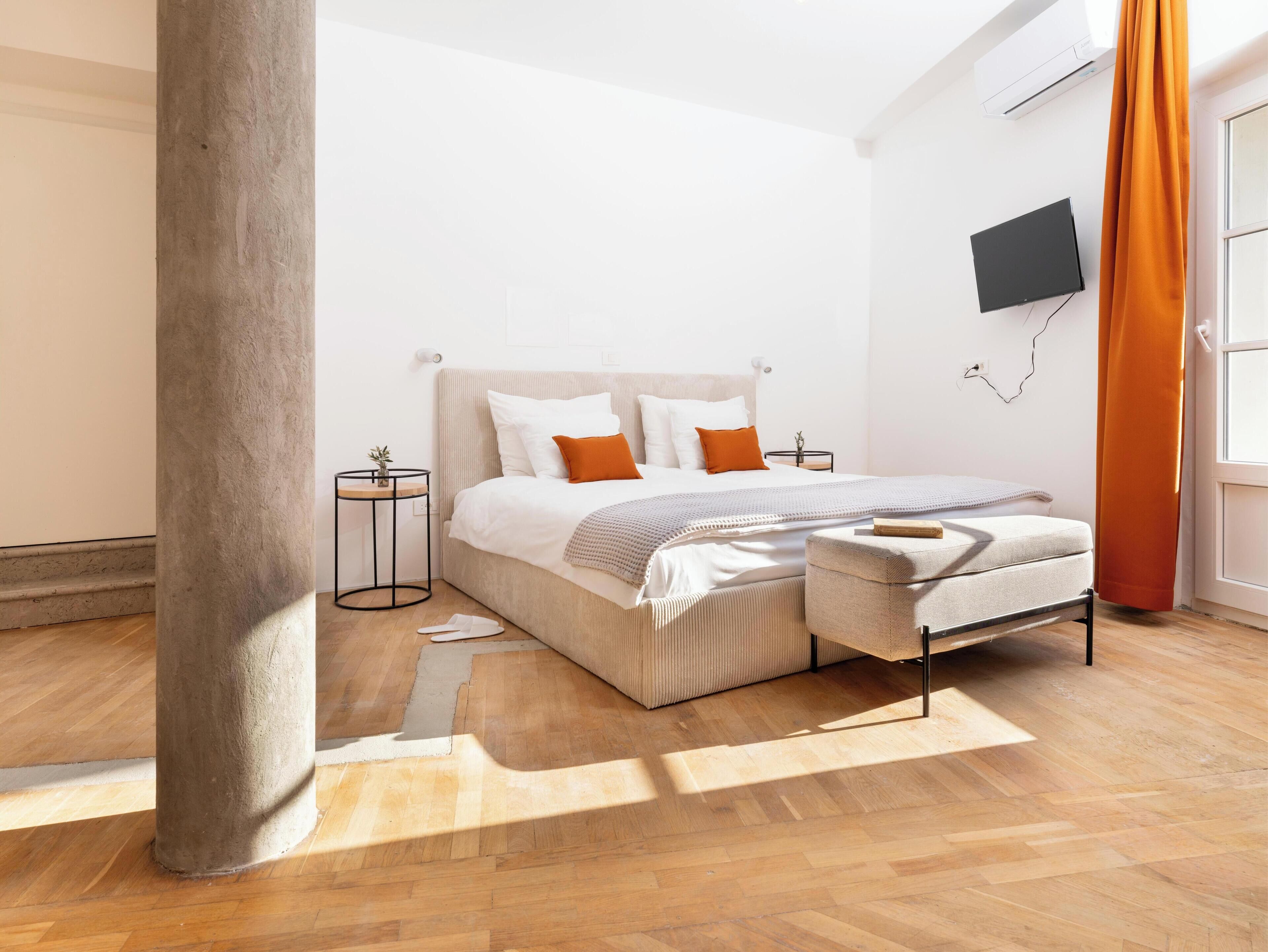 A modern bedroom with a large bed, a small bedside table, a TV mounted on the wall, and a window with curtains.