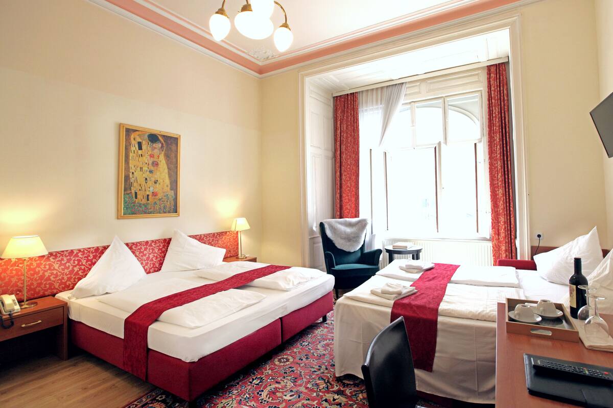 Quarto de hotel com duas camas, uma escrivaninha, uma cadeira, telefone, abajur, pintura e janela com cortinas.