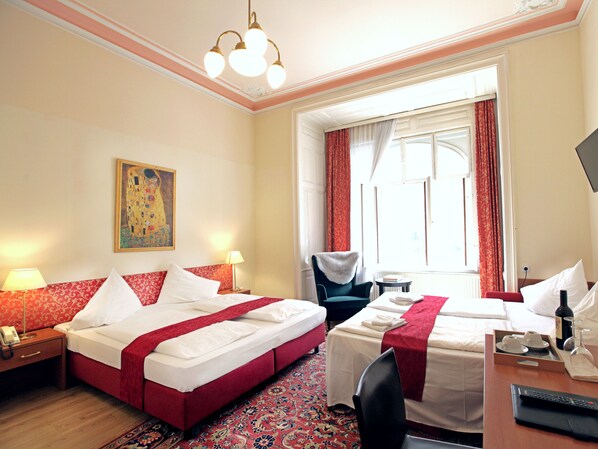 Quarto de hotel com duas camas, uma escrivaninha, uma cadeira, telefone, abajur, pintura e janela com cortinas.
