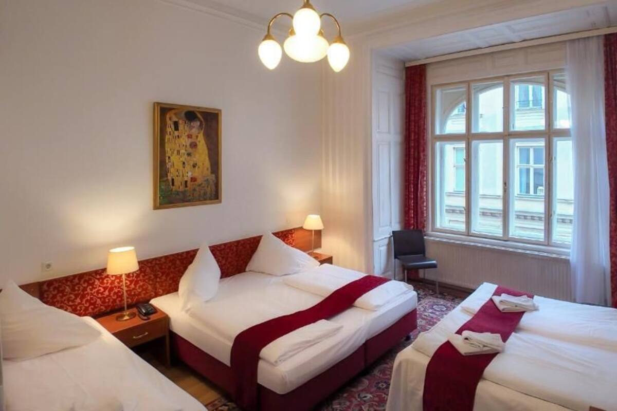 Quarto de hotel com duas camas, uma janela grande com cortinas, um quadro na parede e uma escrivaninha com uma cadeira.