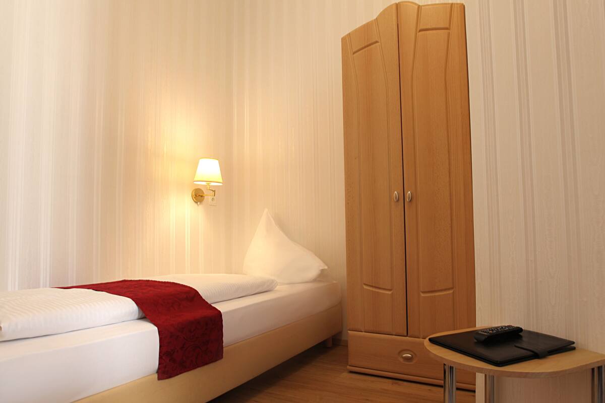 Quarto de hotel com uma cama, um guarda-roupa, uma mesa de cabeceira com controle remoto e uma luminária na parede.