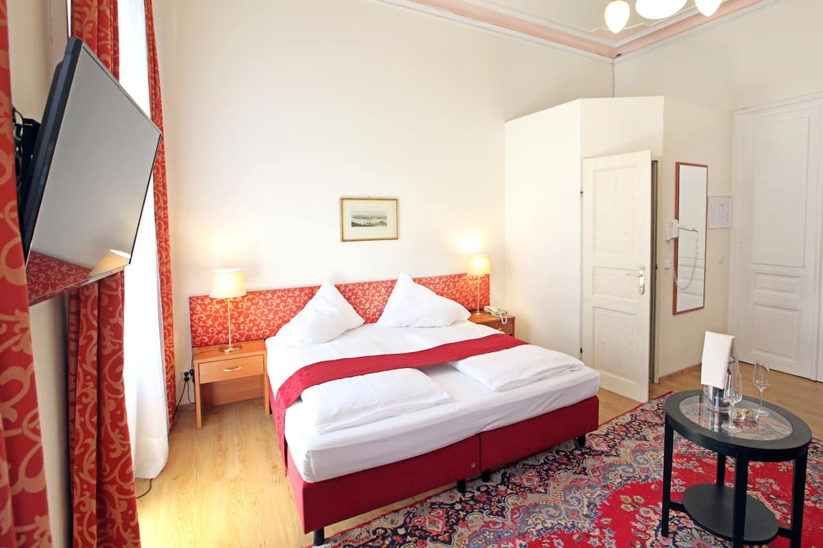 Quarto de hotel com cama, duas luminárias de cabeceira, televisão de tela plana, cabeceira vermelha, espelho grande, porta e pequena mesa com tampo de vidro.