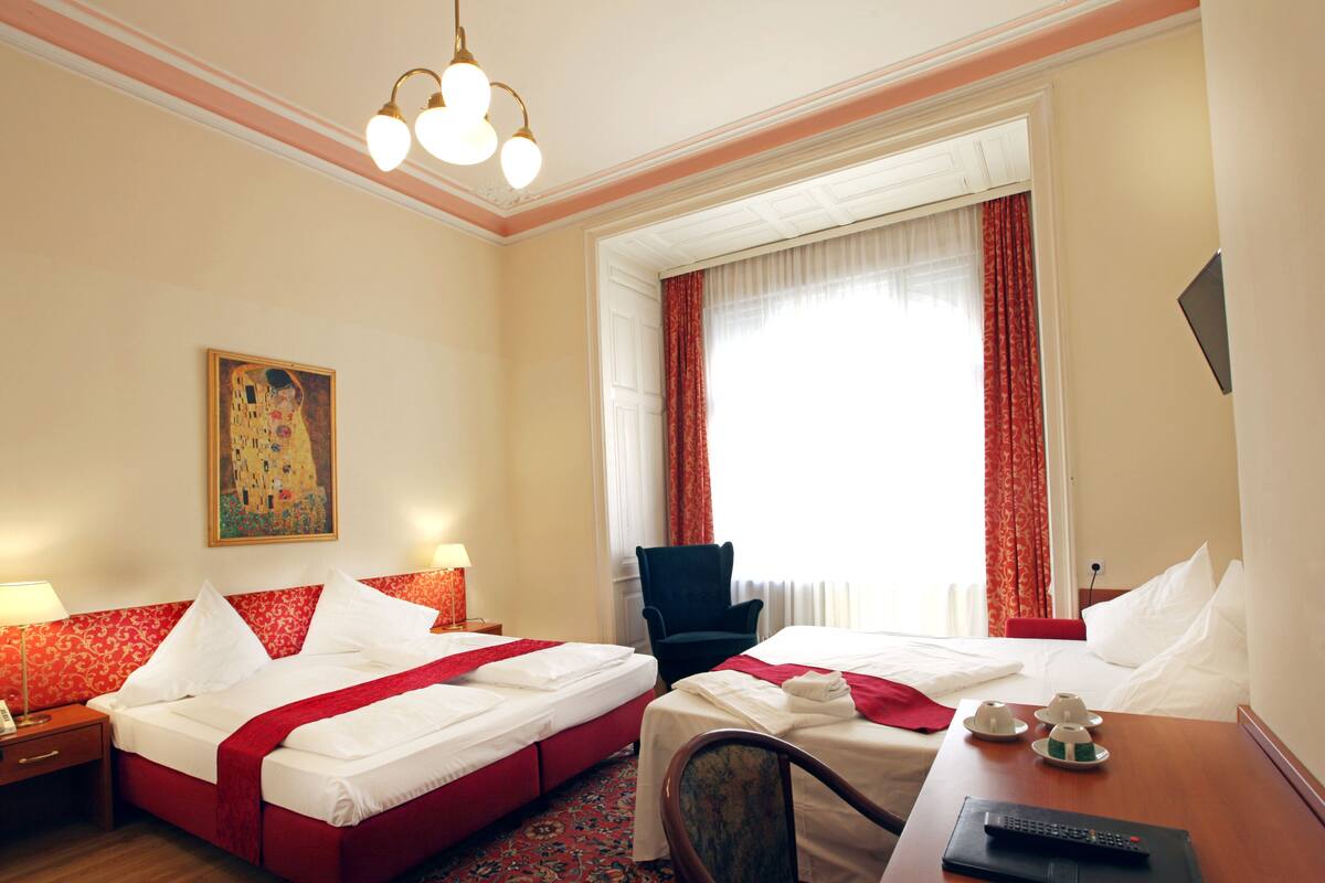 Quarto de hotel com duas camas, uma escrivaninha com controle remoto, uma cadeira, um quadro na parede e uma janela com cortinas.
