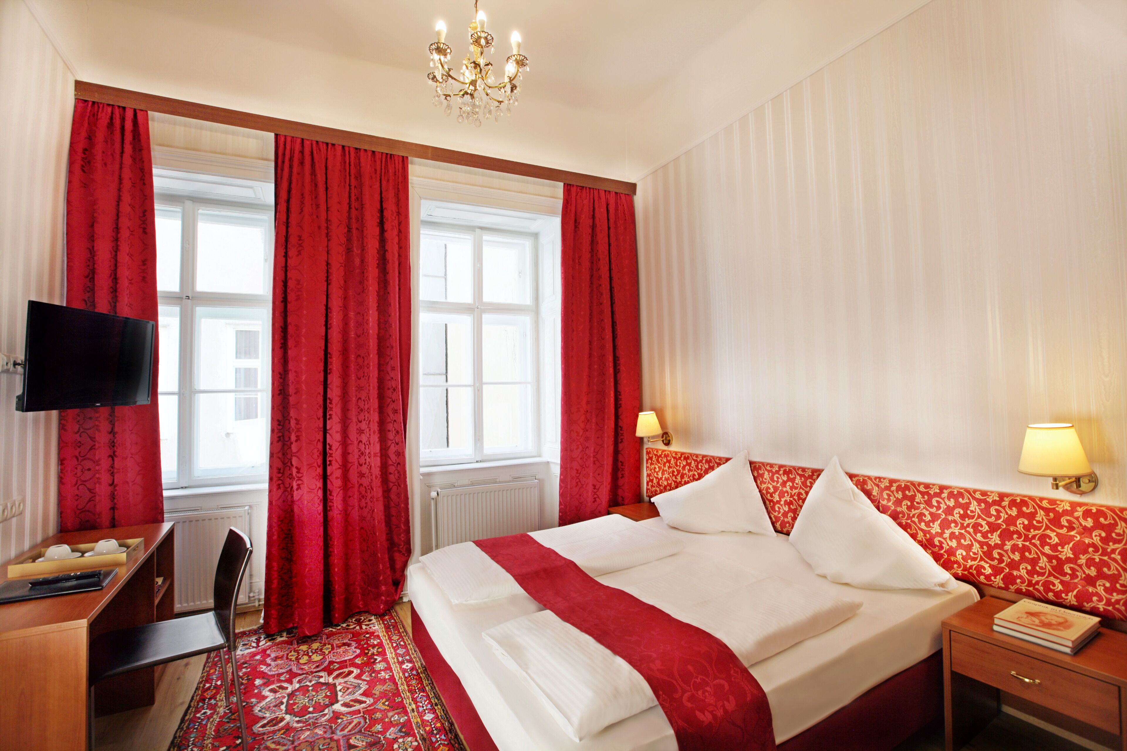 A hotel room with a large bed, red curtains, a television, a desk, and a patterned rug.