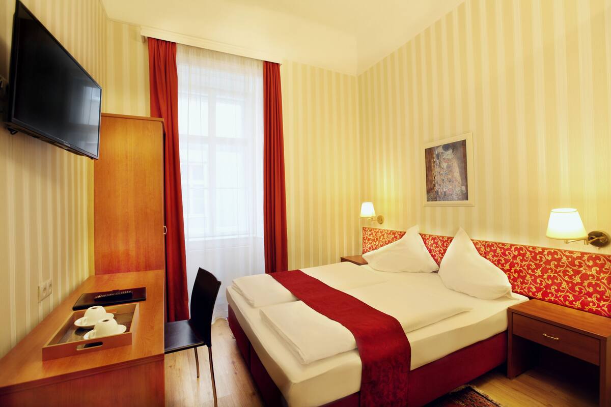 Quarto de hotel com cama, escrivaninha, cadeira, televisão e janela com cortinas.