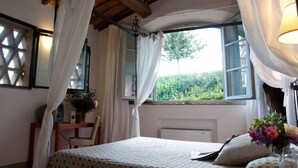 A bedroom with a wooden ceiling, a bed with a patterned bedspread, a bedside table with a lamp, and a window with curtains. - Villa Ducci (San Gimignano)