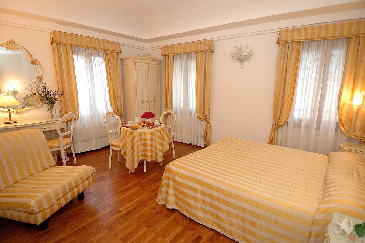 A hotel room with a striped yellow and white sofa, a matching striped bed, a wooden floor, a dining table with chairs, and a large mirror.