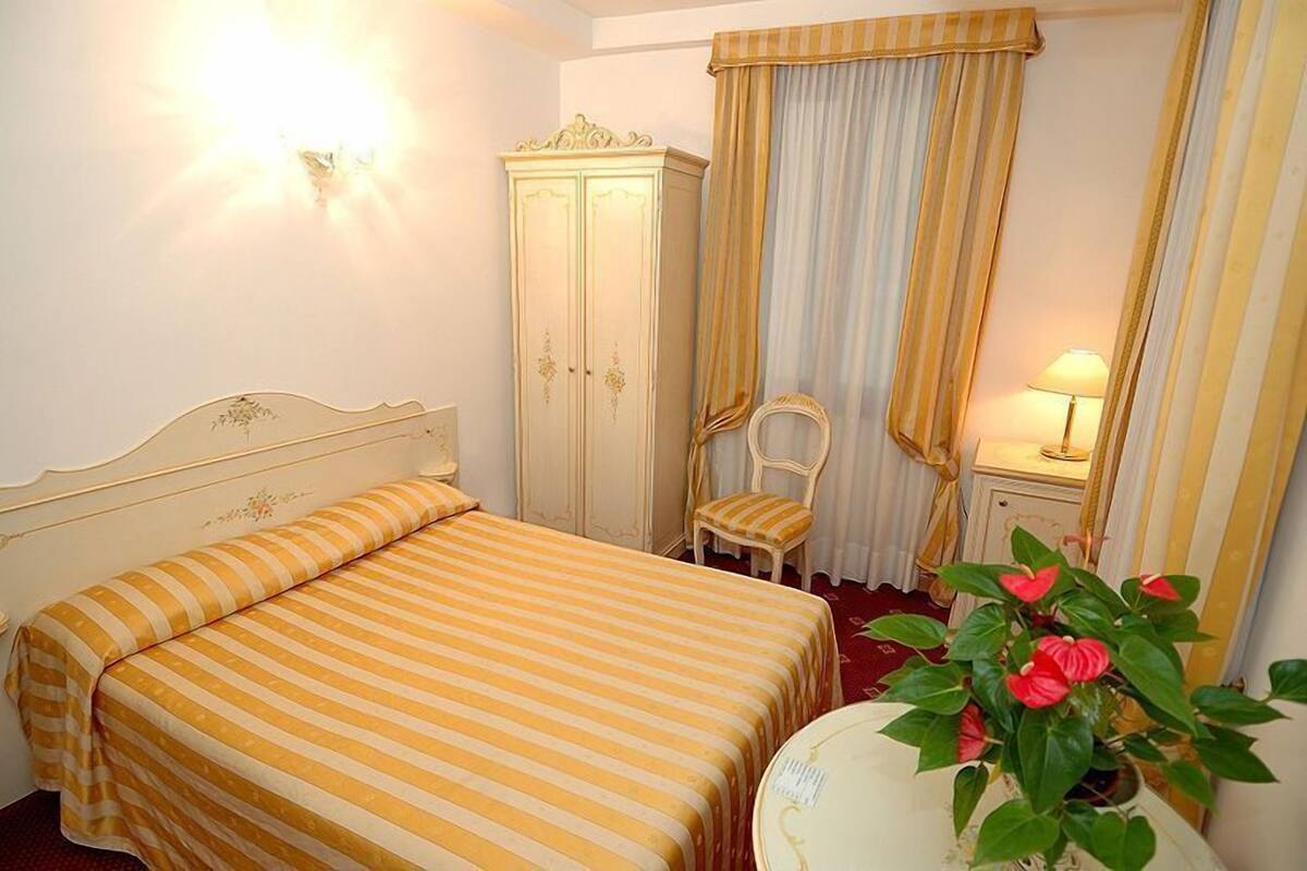 A hotel room with a bed, a chair, a wardrobe, a lamp, and a potted plant.