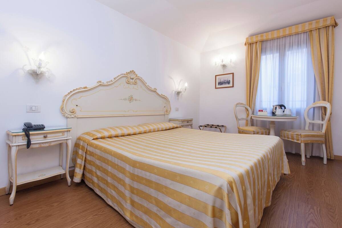 A hotel room with a large bed, a bedside table with a lamp, a chair, a small table with a kettle, and a window with curtains.
